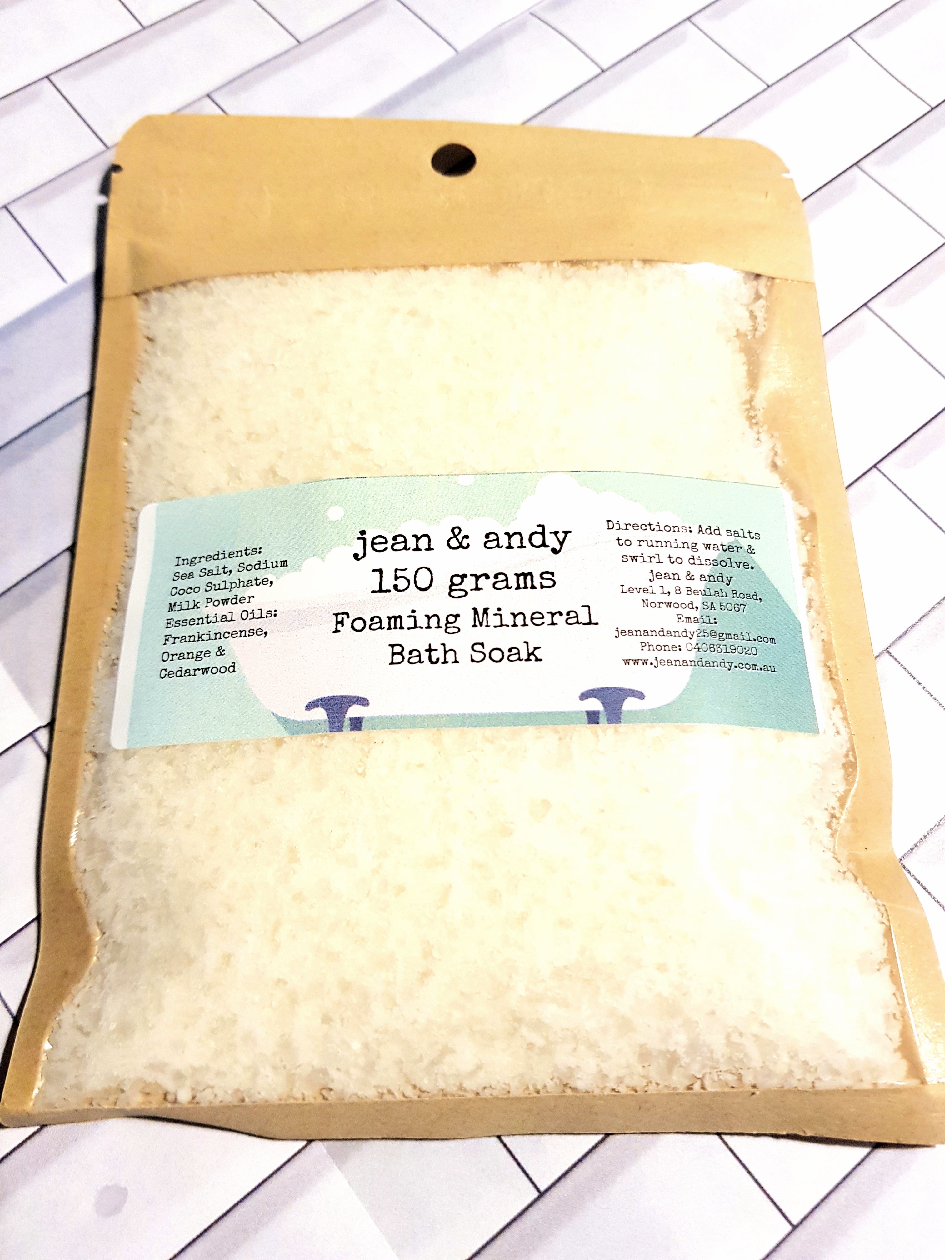 A brown paper package of Foaming Mineral Bath Salts by jean & andy, weighing 150 grams, rests on a white tiled surface. Featuring ingredients such as Epsom salt, coconut milk powder, and essential oils, this luxurious blend promises a rejuvenating experience.