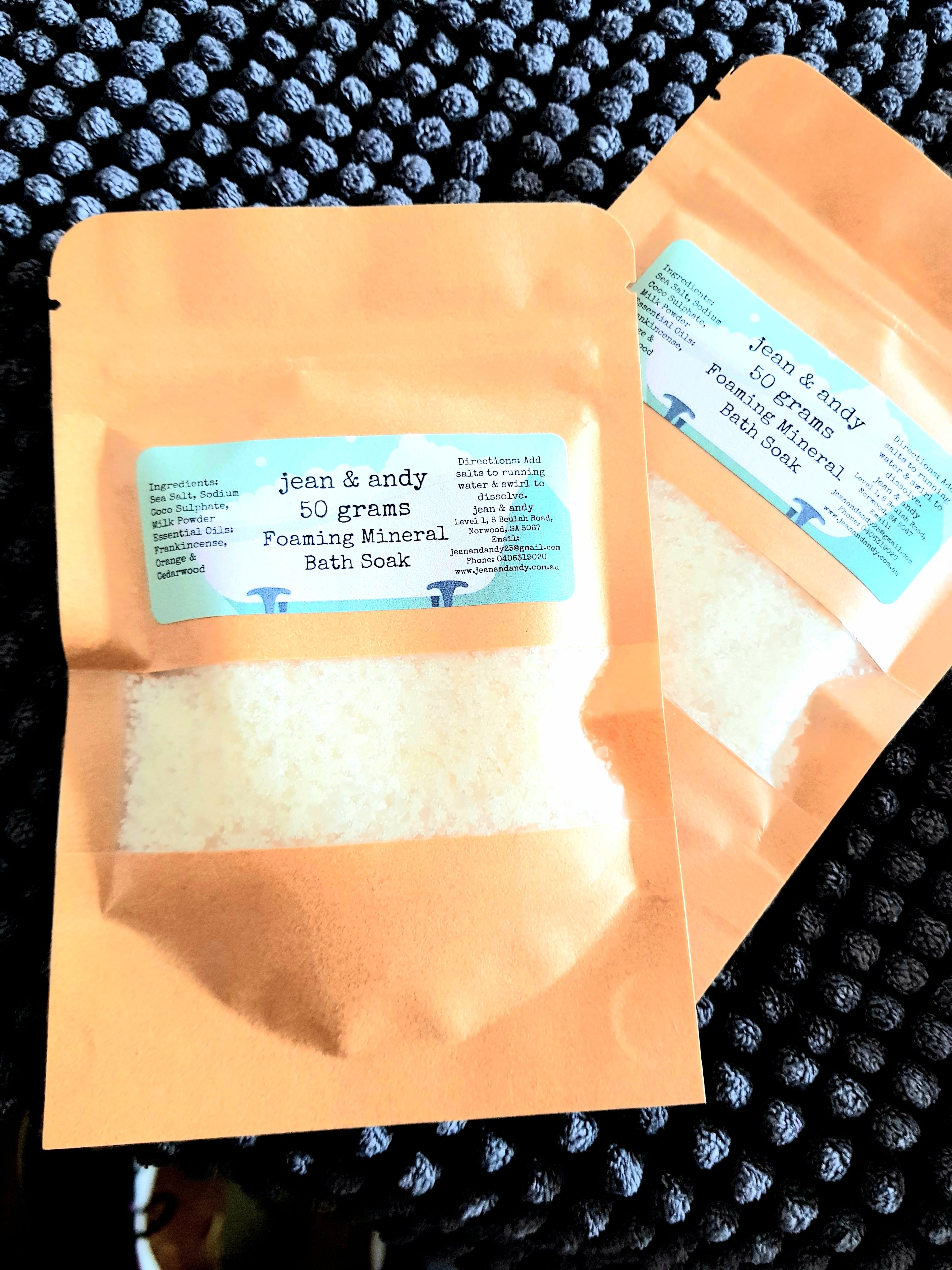 Two brown paper pouches of Foaming Mineral Bath Salts by jean & andy, each containing 50 grams and enriched with sea salt and calming essential oils, elegantly rest on a textured dark surface.