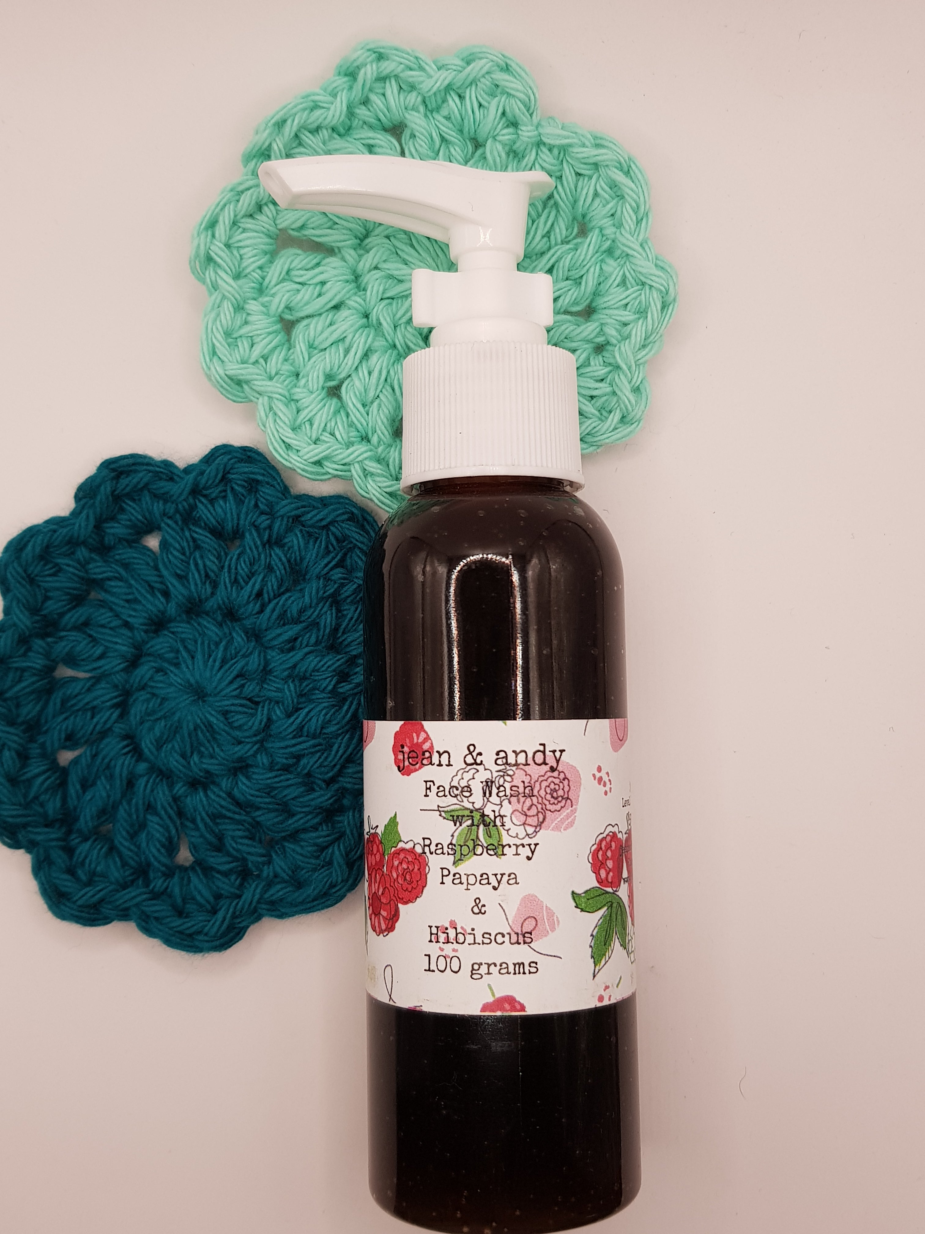 A bottle of Raspberry Papaya & Hibiscus face wash is accompanied by two jean & andy Face Scrubbies, available in teal and mint green, set against a white background. This eco-conscious arrangement seamlessly blends style with environmentally friendly skincare.