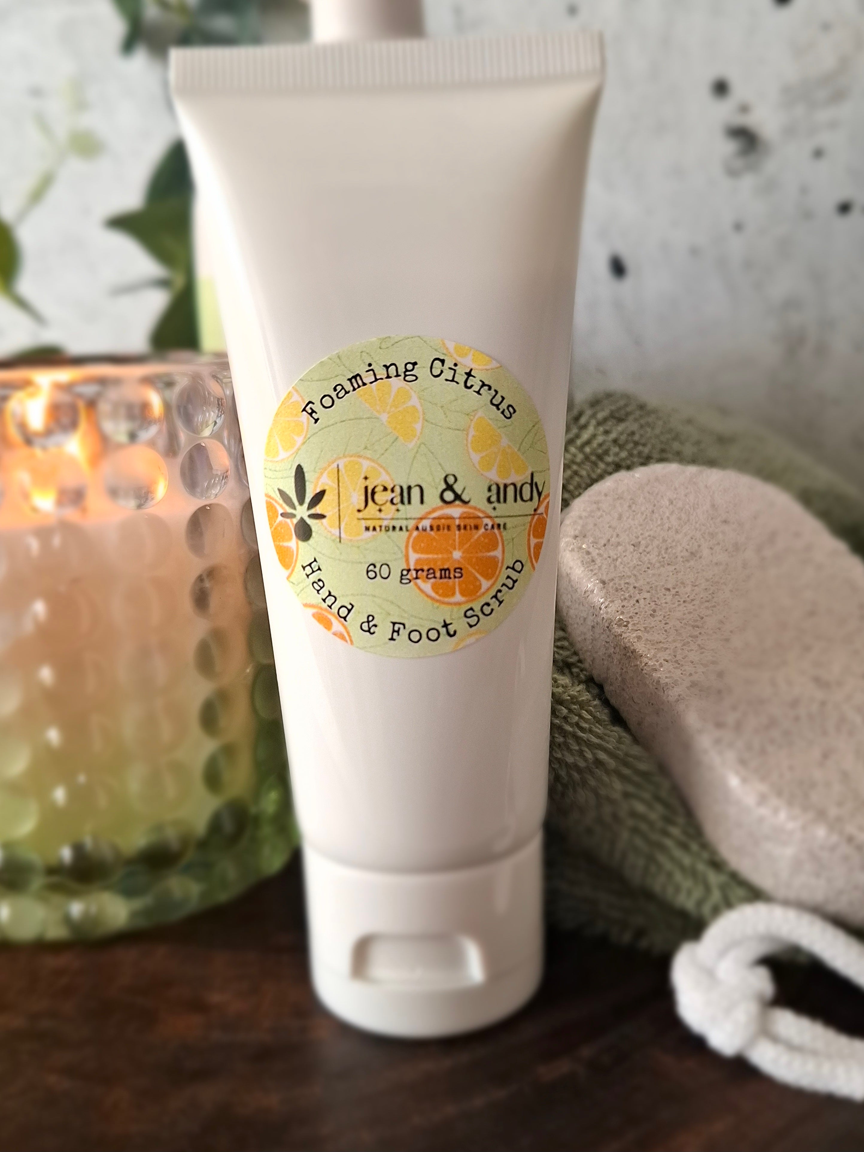 A tube of jean & andy Foaming Citrus Hand & Foot Scrub, infused with essential oils for a luxurious exfoliation, rests on a table next to a lit candle, a pumice stone, and a green cloth, creating a relaxing spa-like atmosphere.