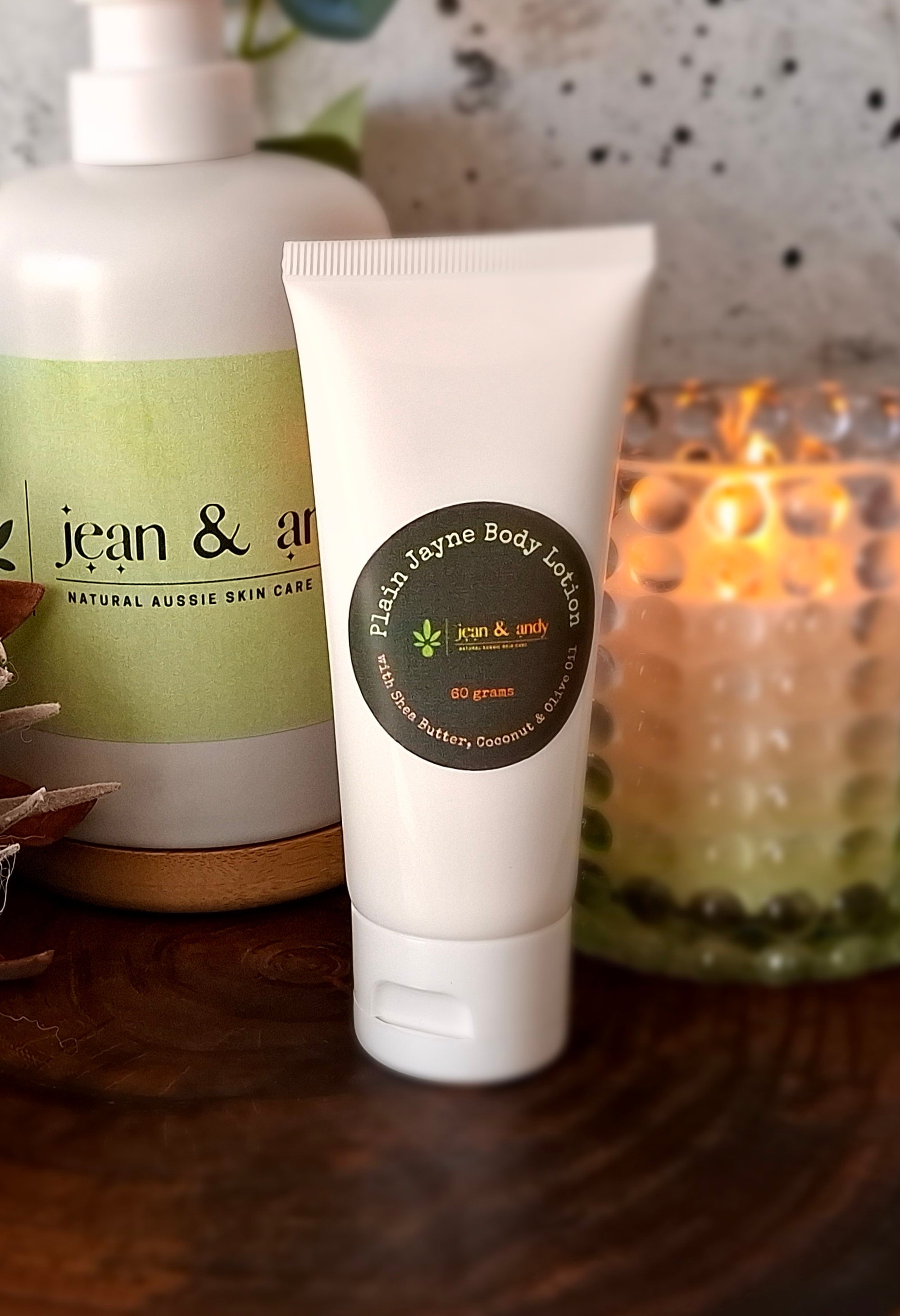 A tube of jean & andy Plain Jayne Body Lotion stands on a counter. In the background, there's a larger bottle and a lit candle, enhancing the warm ambiance with its soothing light. The lotion label mentions coconut and honey for skin hydration, offering antioxidant protection for your daily skincare routine.