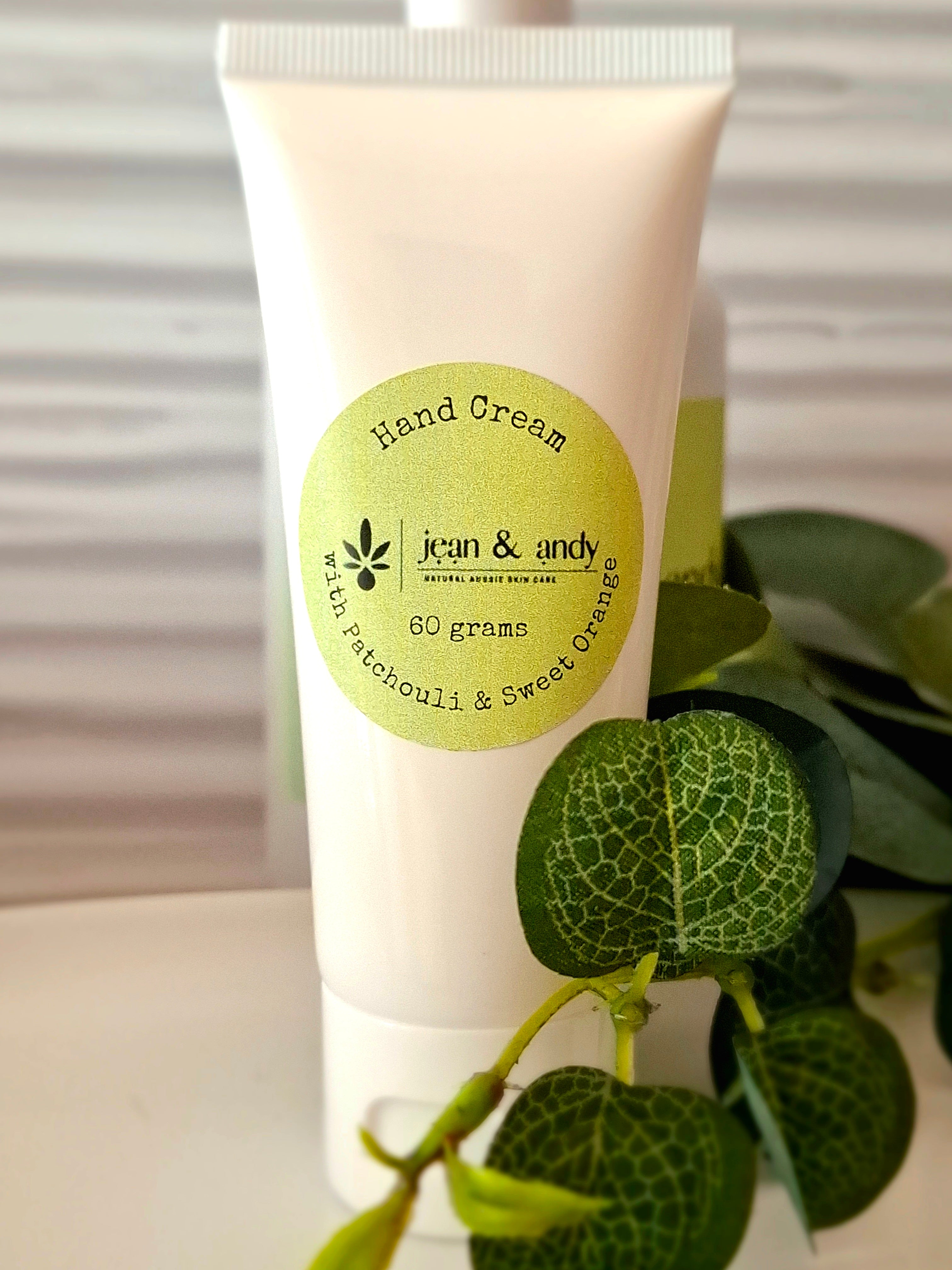 A beige tube of jean & andy's "Patchouli and Sweet Orange Hand Cream," featuring a green label, weighs 60 grams and is infused with Patchouli Oil and Sweet Orange essence. Green leafy branches in the background enhance its presentation.