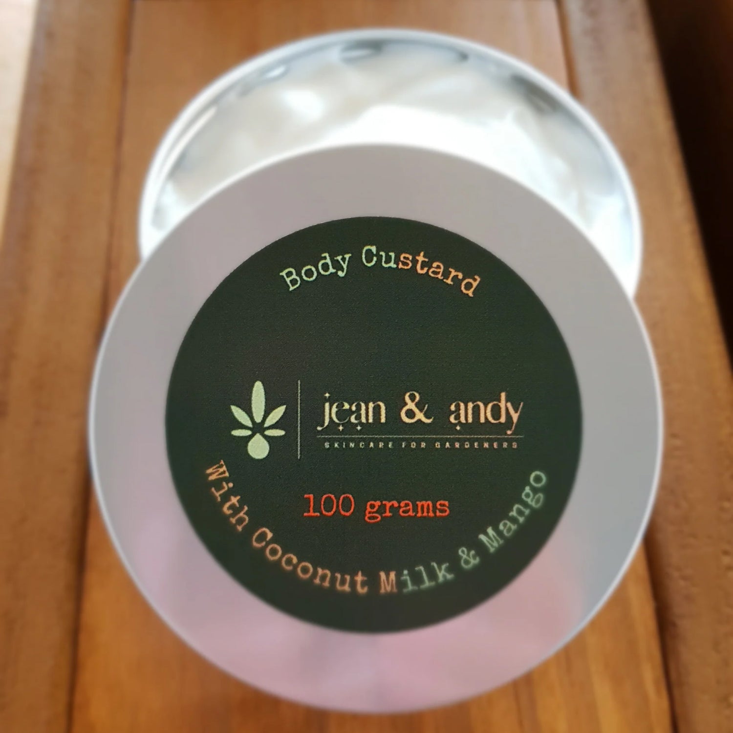 A round silver tin labeled Body Custard with Coconut & Mango by jean & andy is displayed, containing 100 grams of a luxurious blend. The tin rests gracefully on a wooden surface, its lid slightly open to reveal the tropical infusion within.