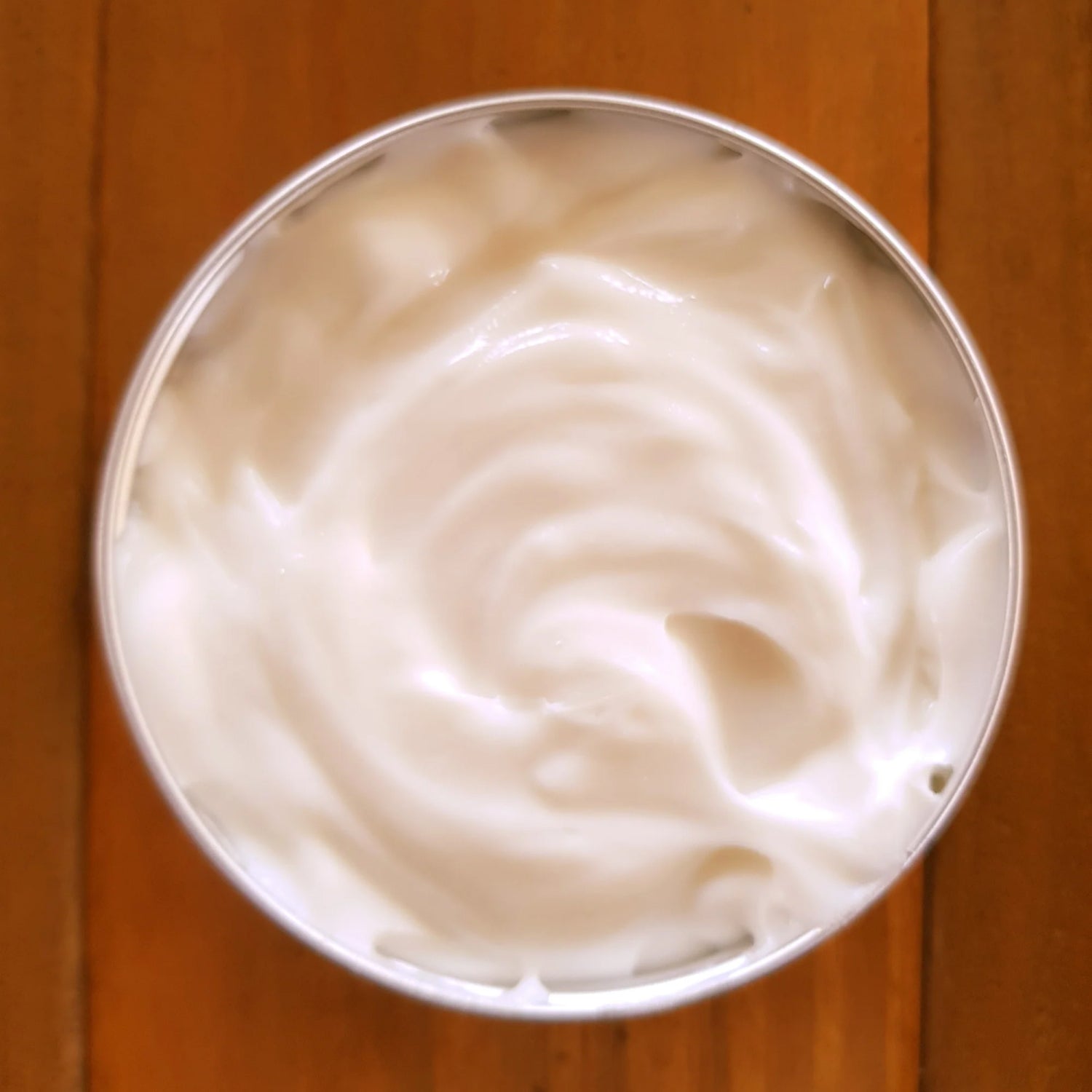 A round, open tin of jean & andy's Body Custard with Coconut & Mango rests on a wooden surface, revealing its smooth, creamy white substance infused with the essence of coconut milk. The custard showcases a soft, swirling texture.