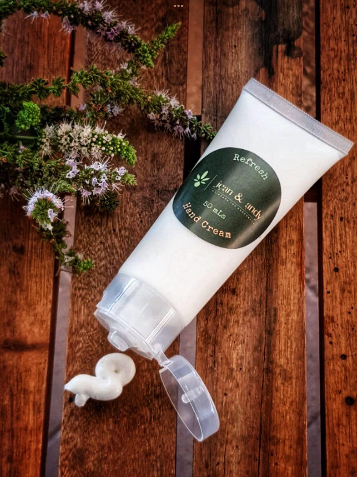 A tube of jean & andy's Refresh Hand Cream sits on a wooden surface, with a swirl of its rich formula containing essential oils squeezed out. A sprig of green leaves and tiny purple flowers enhances the peppermint-infused scene, bringing a natural touch.