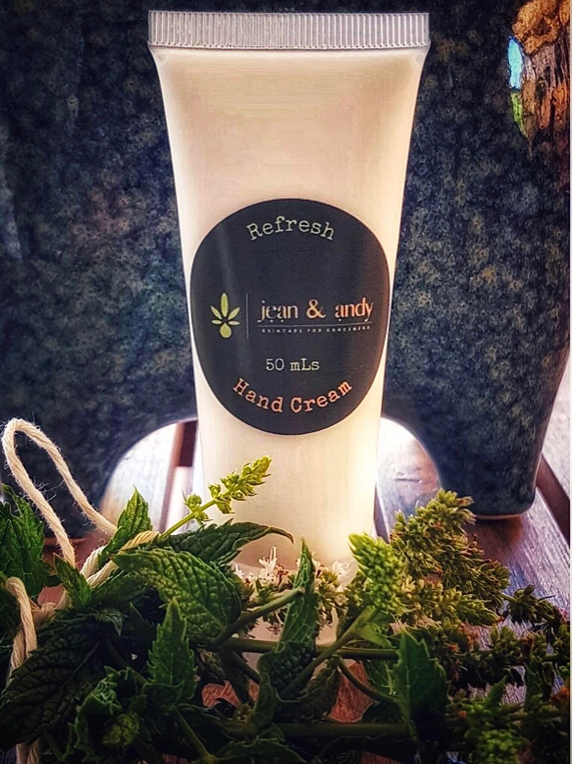 A 50 mL tube of jean & andy's Refresh Hand Cream, enriched with essential oils, is elegantly presented on a wooden surface accompanied by a small bouquet of mint leaves. A textured ceramic vase serves as the background.