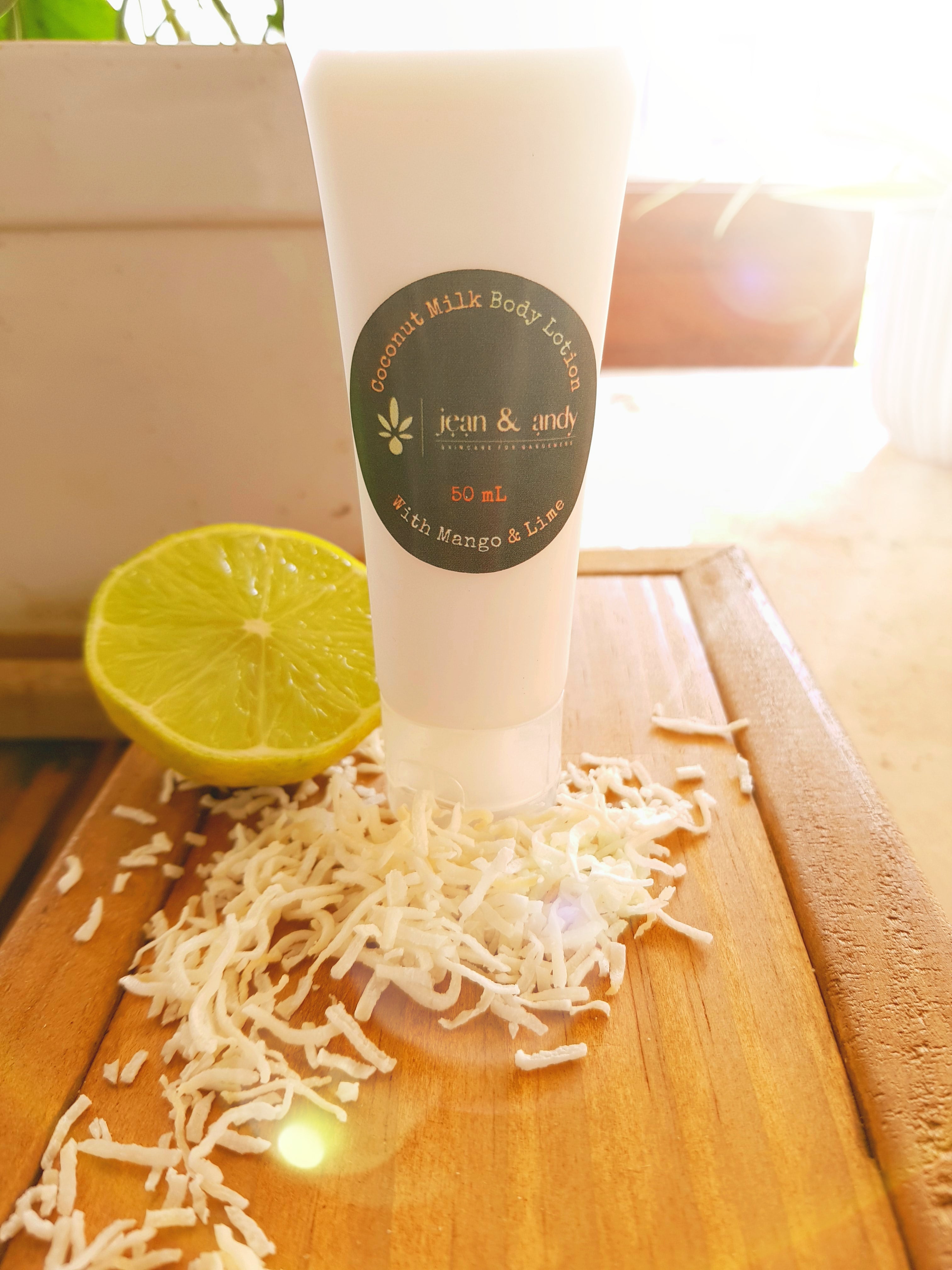 The Coconut Milk Body Lotion by jean & andy, enriched with shea butter, is showcased alongside shredded coconut and a halved lime on a wooden surface, illuminated by streaming sunlight.