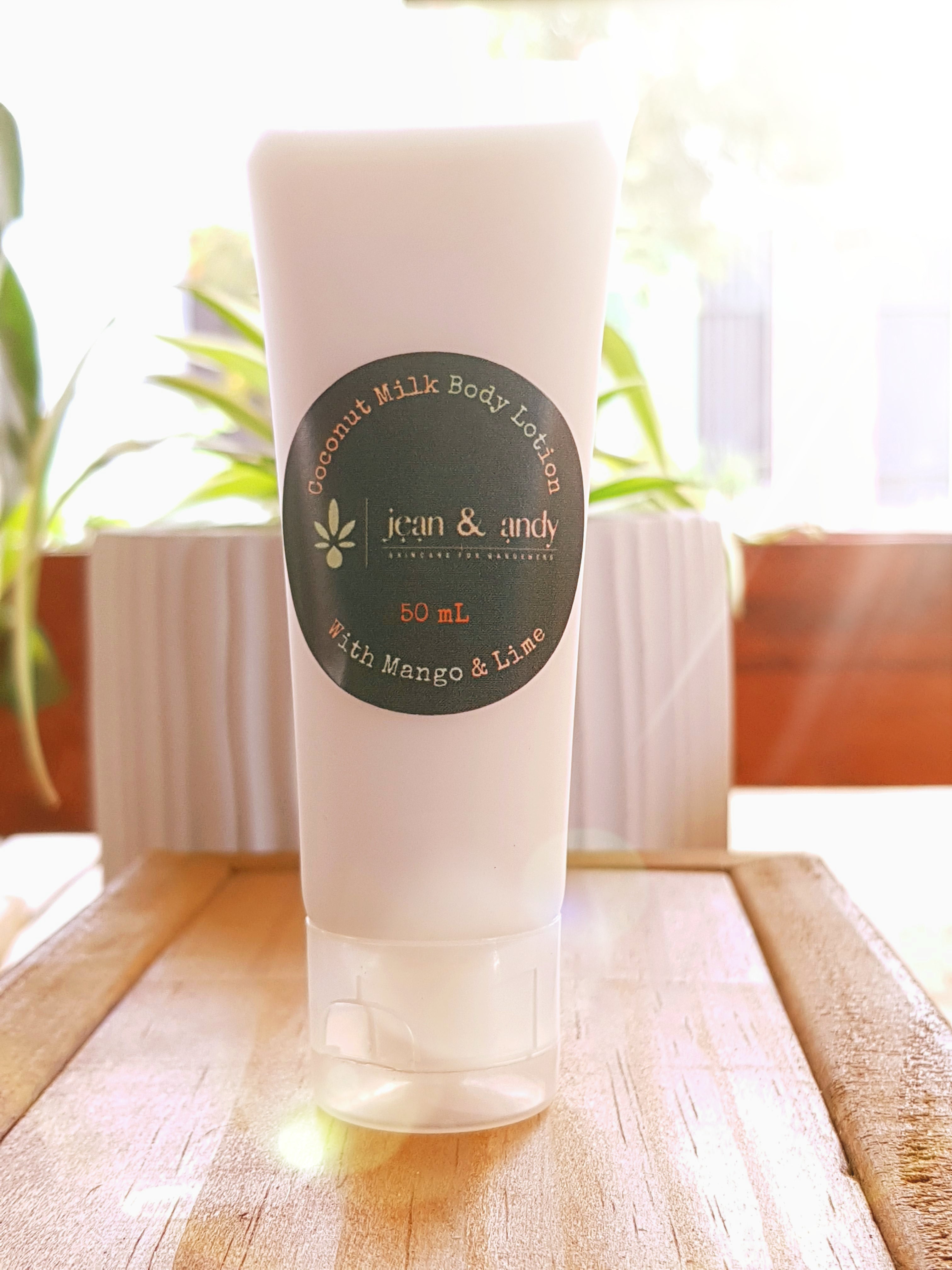 A softly-lit image showcases a white 50mL tube of Coconut Milk Body Lotion, adorned with jean & andy's green and gold logo. The lotion highlights ingredients like coconut milk powder, mango, and lime. It is placed on a wooden surface with sunlight streaming in and greenery softly blurred in the background.
