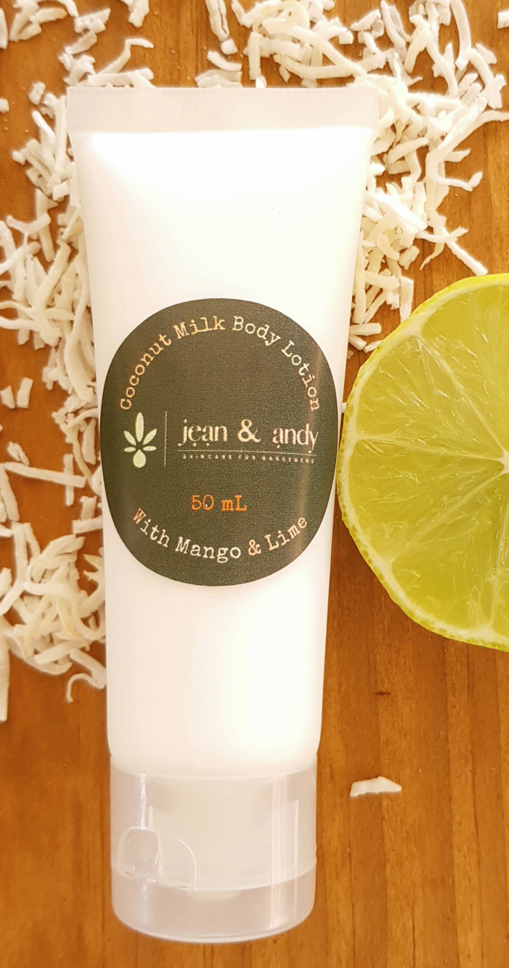 A tube of jean & andy Coconut Milk Body Lotion with a mango and lime aroma rests on a wooden surface, encompassed by shredded coconut and a slice of lime. Enriched with shea butter for additional nourishment, it provides a tropical escape in every use.