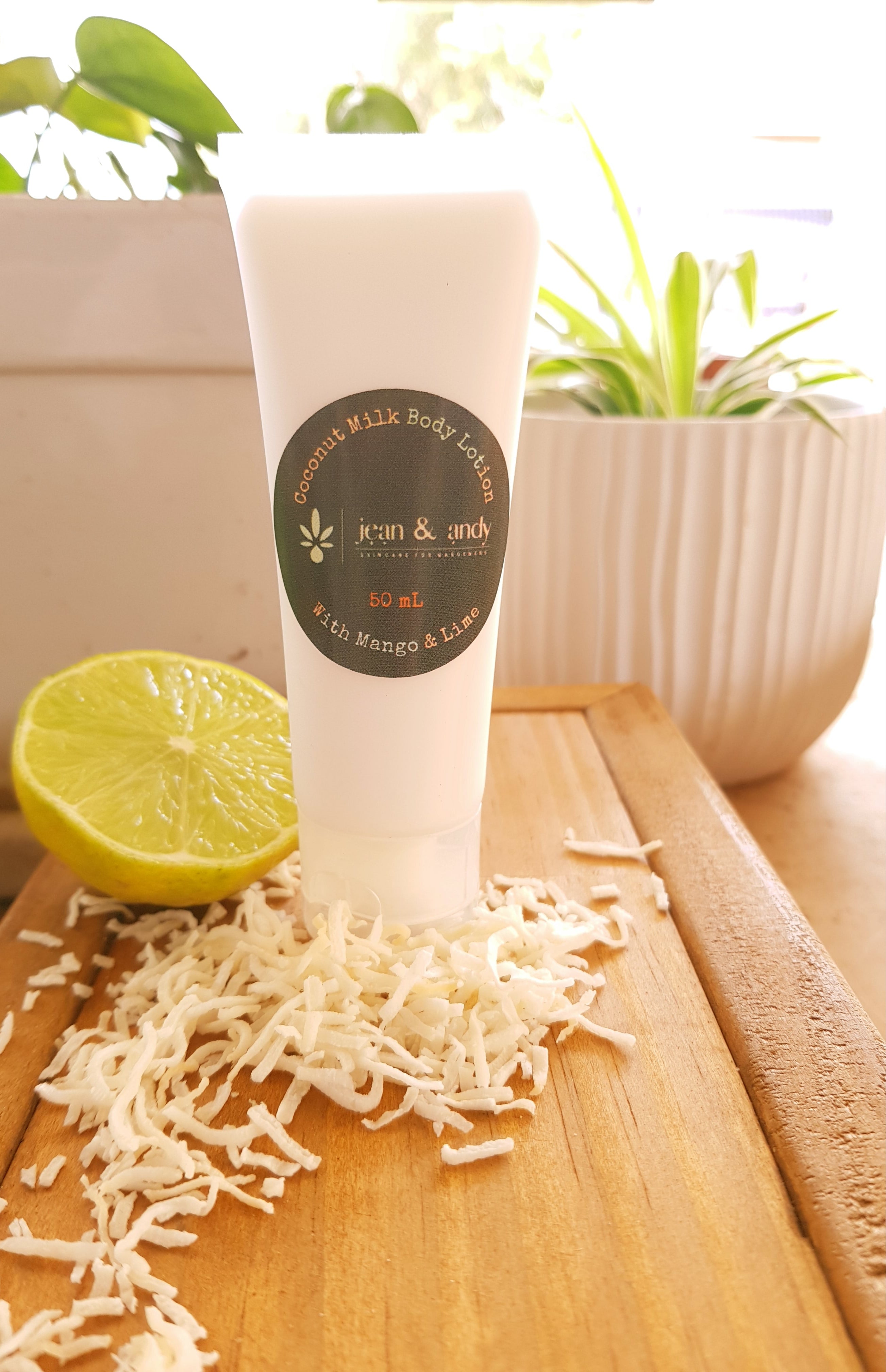 A tube of Coconut Milk Body Lotion from jean & andy, enriched with nourishing shea butter, rests on a wooden board surrounded by shredded coconut. Nearby, a halved lime complements the scene, while a potted plant in the background adds a touch of greenery.