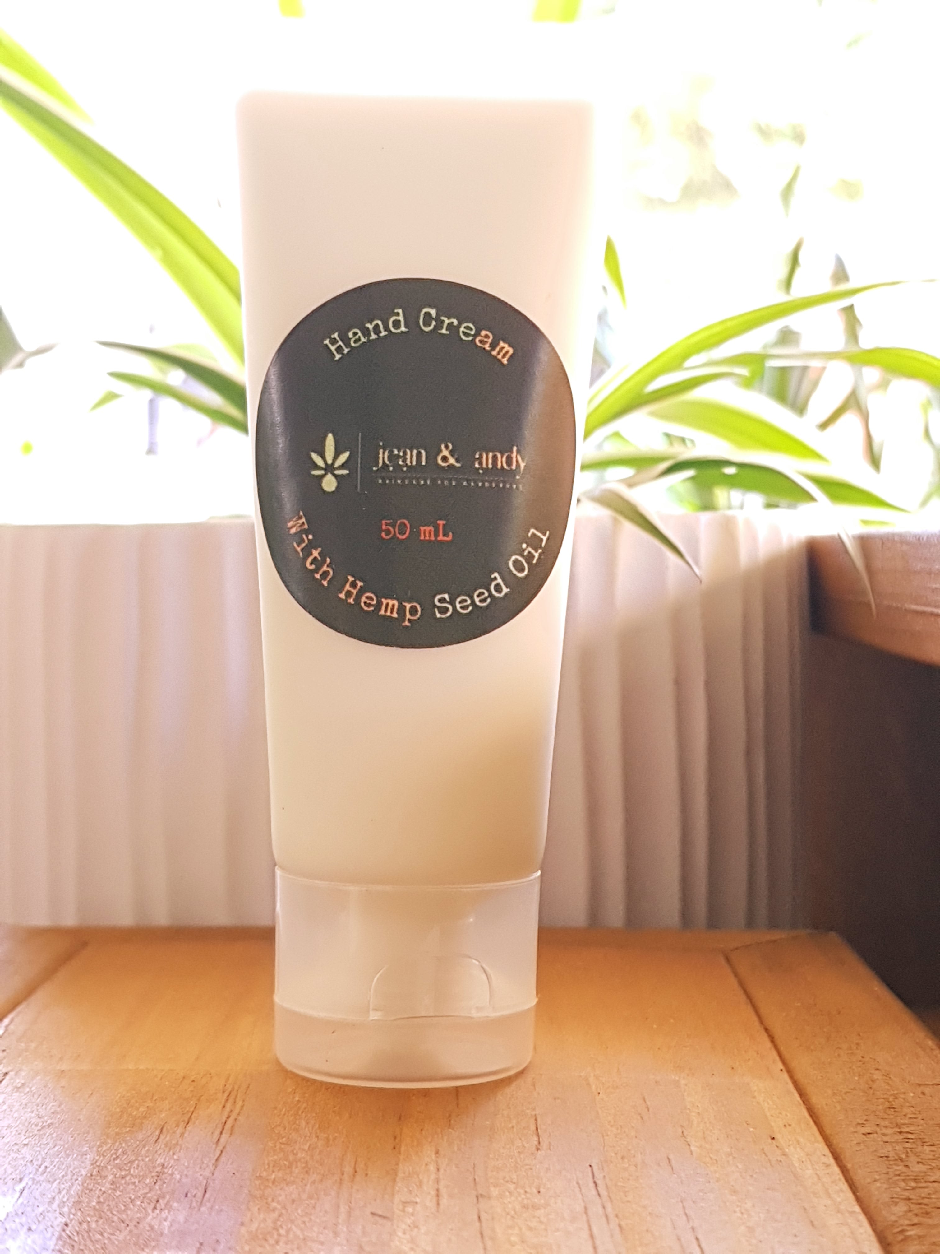 A 50 mL tube of Hand Cream with Hemp Seed Oil by jean & andy, ideal for sensitive skin, is beautifully captured on a wooden surface amidst sunlight and lush plants, highlighting its vegan-friendly properties.