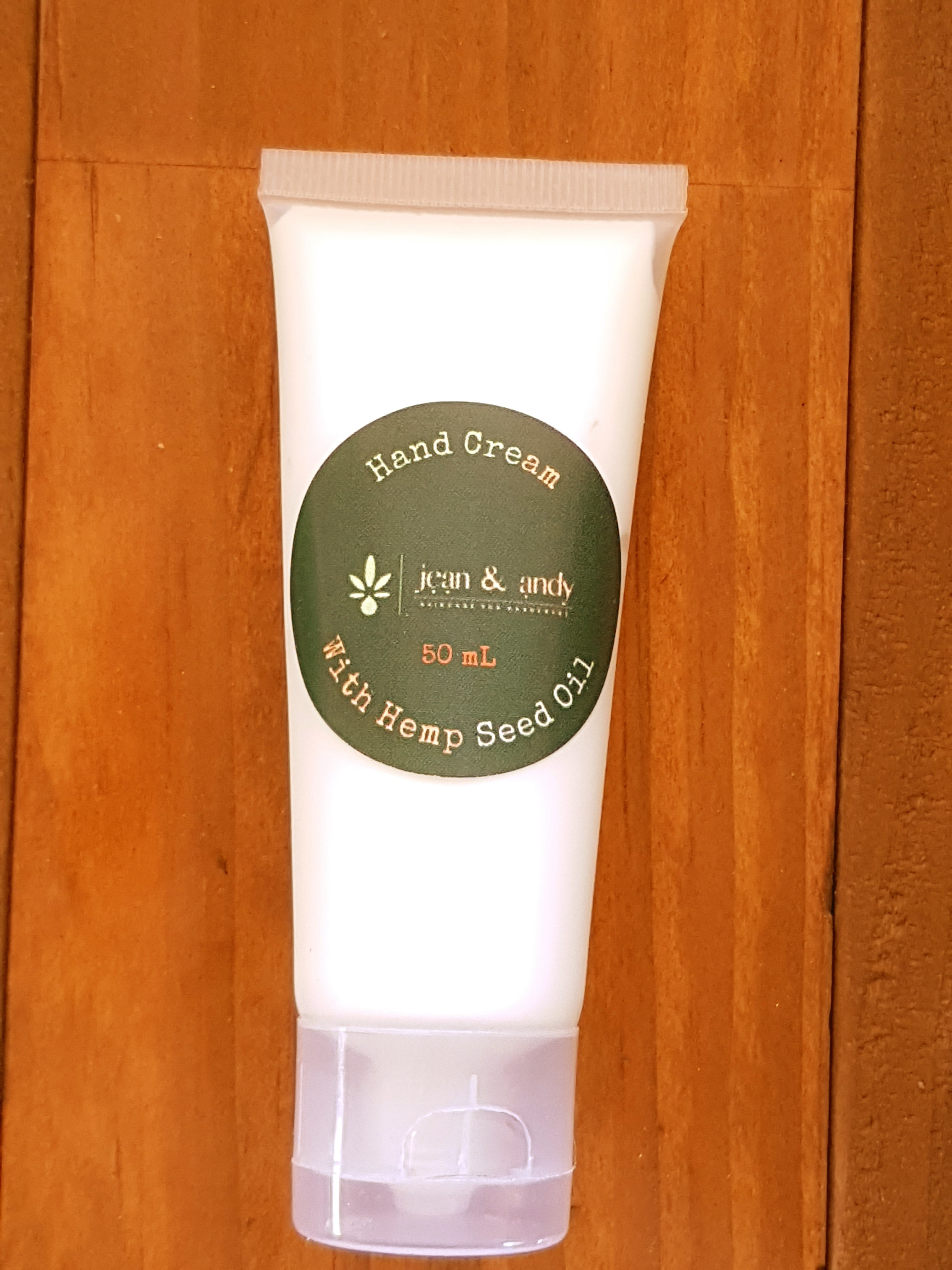 A white tube of hand cream featuring a green circular label that says Hand Cream with Hemp Seed Oil, jean & andy, 50 mL, stands upright on a wooden surface. Ideal for sensitive skin, this vegan-friendly formula nourishes and hydrates.