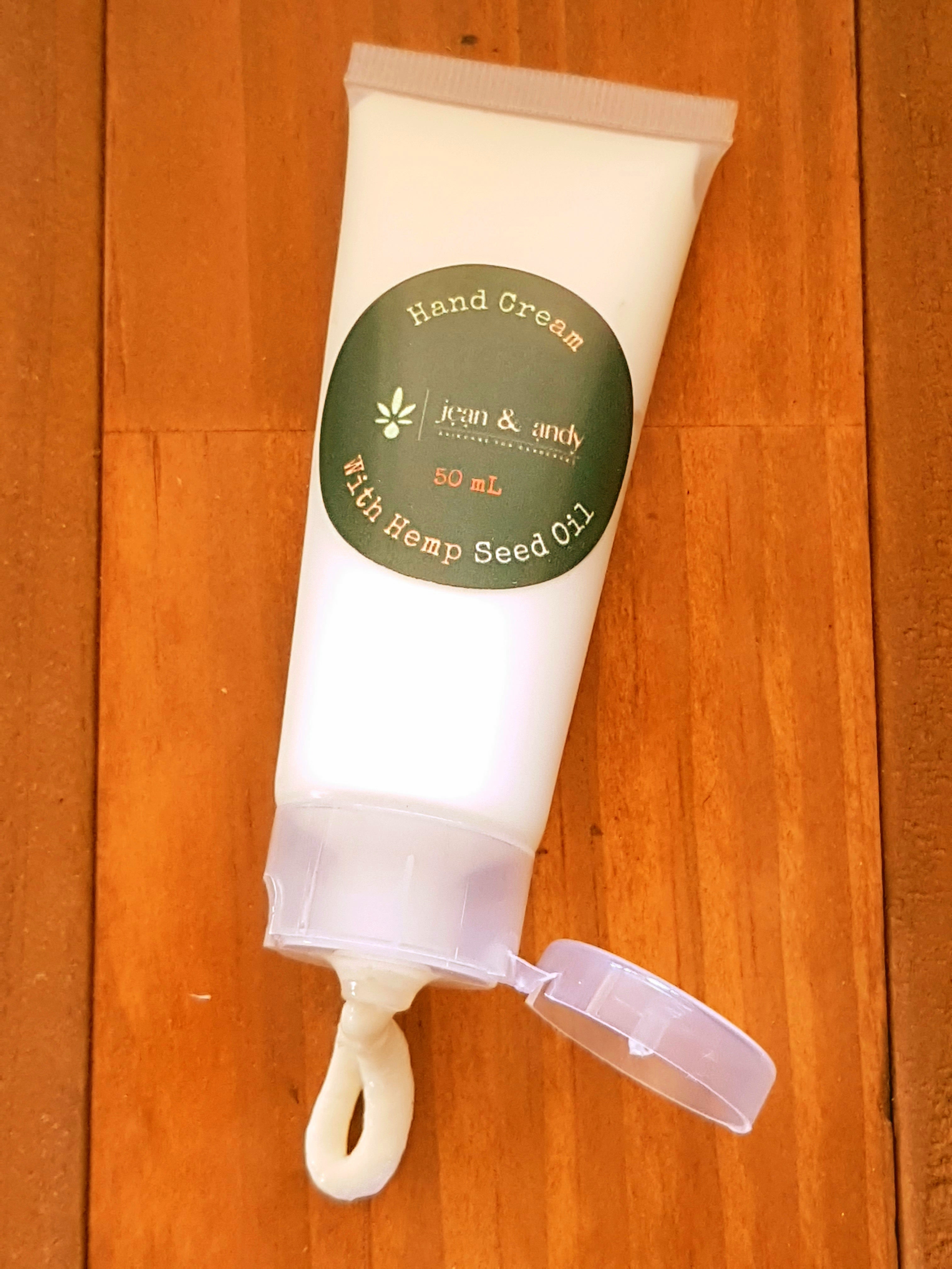 A tube of jean & andy's Hand Cream with Hemp Seed Oil rests on a wooden surface. The green label with white text and a small leaf design indicates its suitability for sensitive skin. The cap is open, revealing a small amount of cream at the opening.