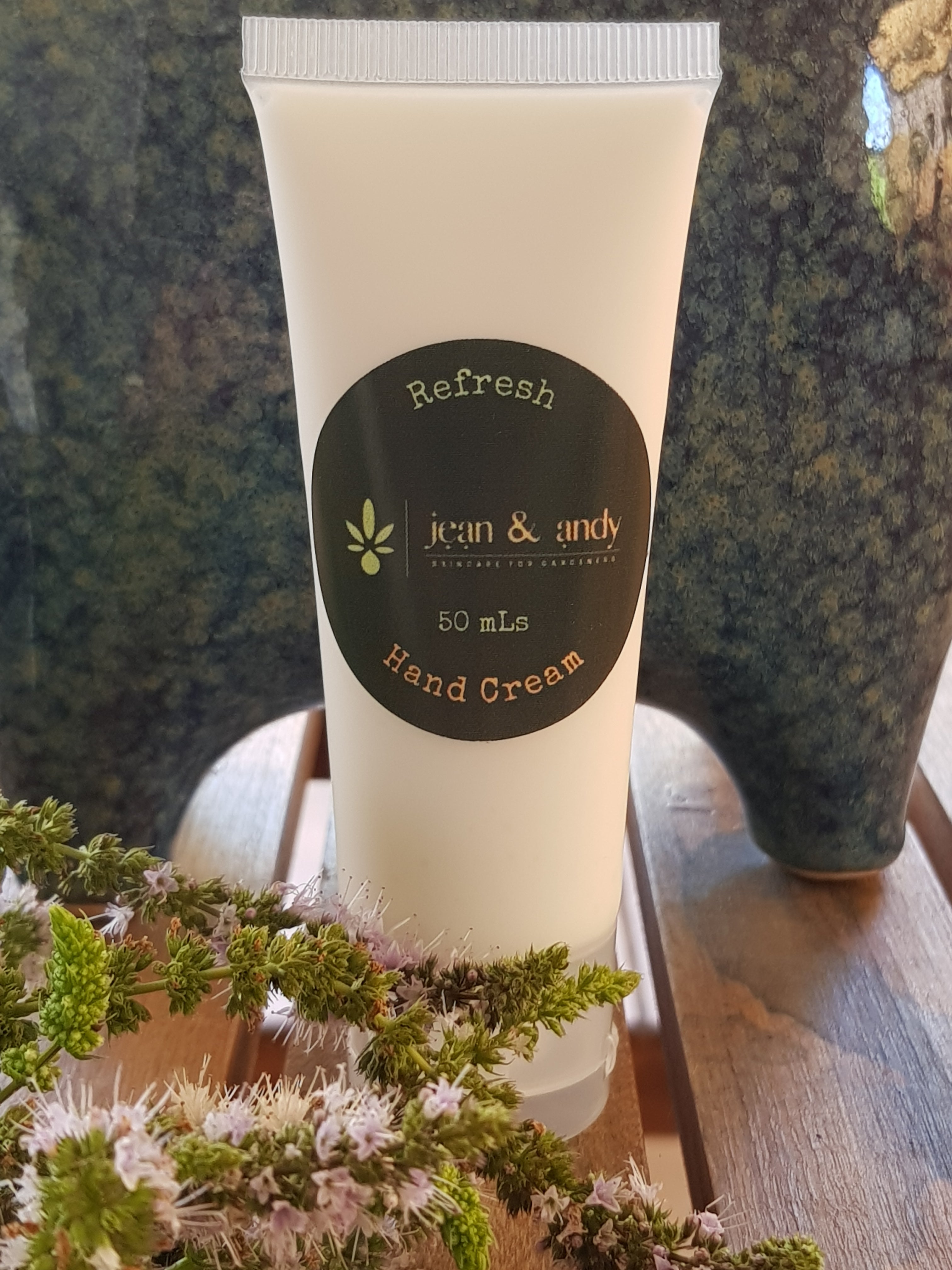 A tube of Refresh Hand Cream by jean & andy, infused with essential oils for extra hydration, sits atop a wooden surface. It has a black label adorned with green decorative details. Close by is a sprig adorned with small flowers, while partially visible textured ceramic items can be seen in the background.