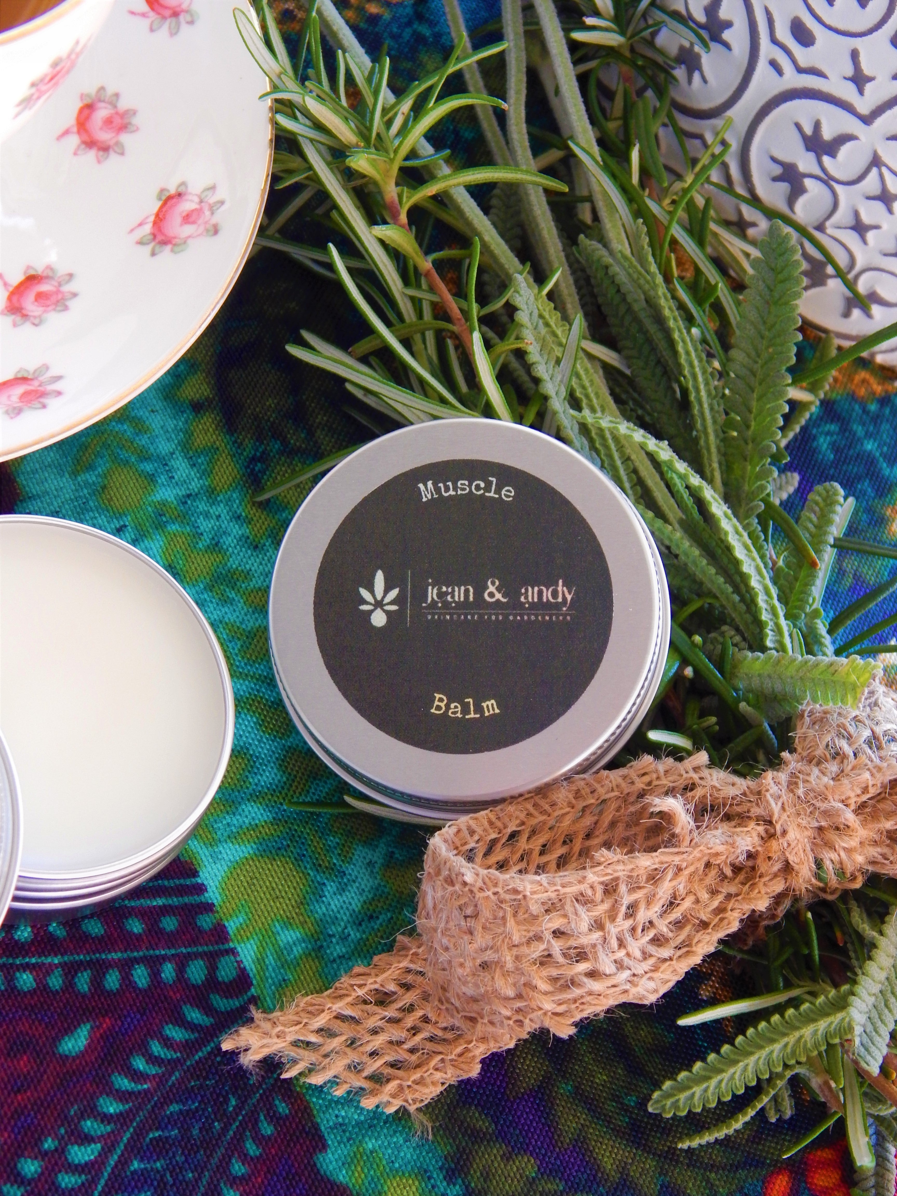 A tin labeled Muscle Balm from jean & andy sits open on a colorful cloth. Nearby are sprigs of rosemary and lavender, rich in essential oils, accompanied by a floral teacup and burlap ribbon, creating a natural, soothing arrangement with an analgesic effect.