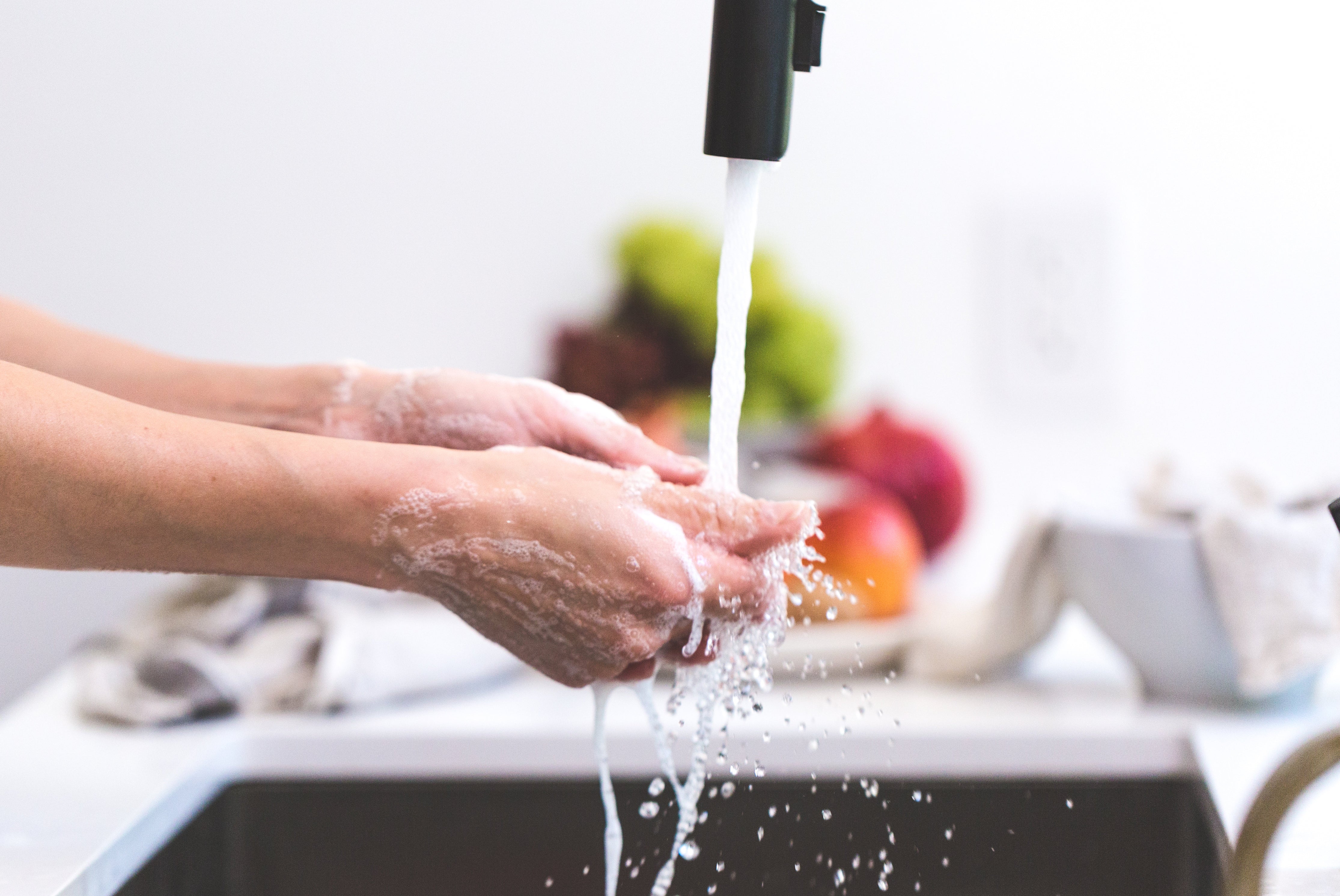 A close-up view shows hands being washed under running water in a kitchen sink. The Foaming Citrus Hand & Foot Scrub by jean & andy creates a rich lather on the skin, offering gentle exfoliation as the water splashes around. In the background, blurred fruits and a light-colored cloth are visible.