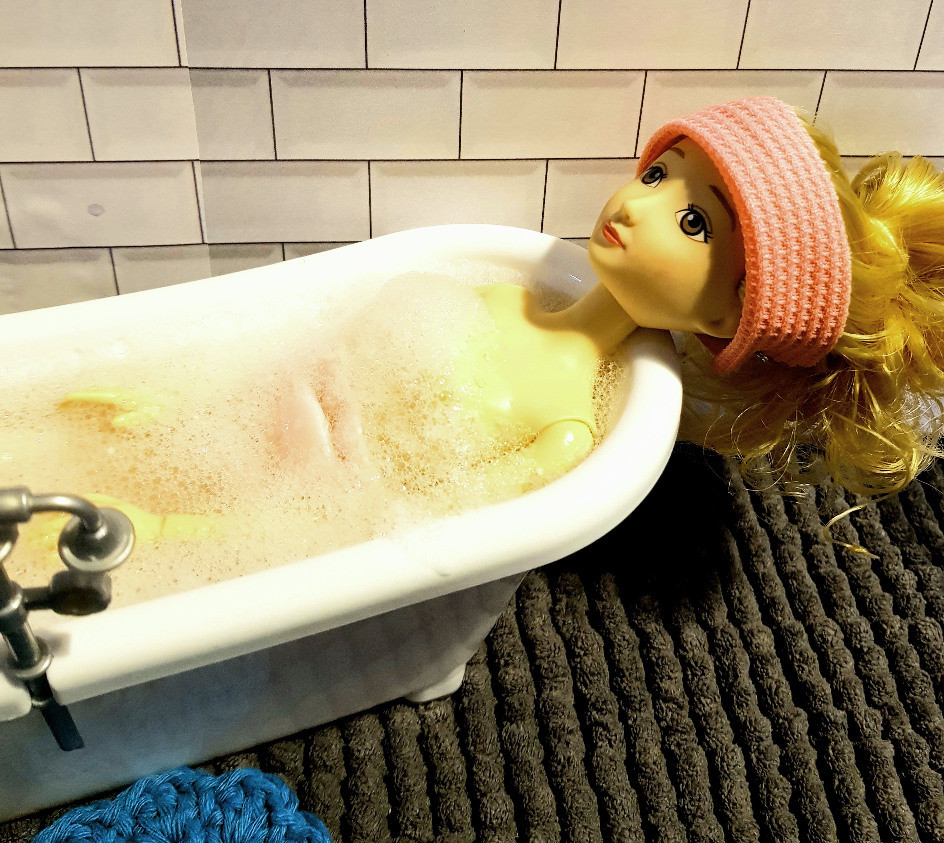 A doll with blonde hair is unwinding in a bubble-filled bathtub, enhanced by jean & andy's Foaming Mineral Bath Salts, while donning a pink headband. The bathtub is positioned against a tiled wall, and the floor features a textured dark mat with a blue bath sponge resting on it.