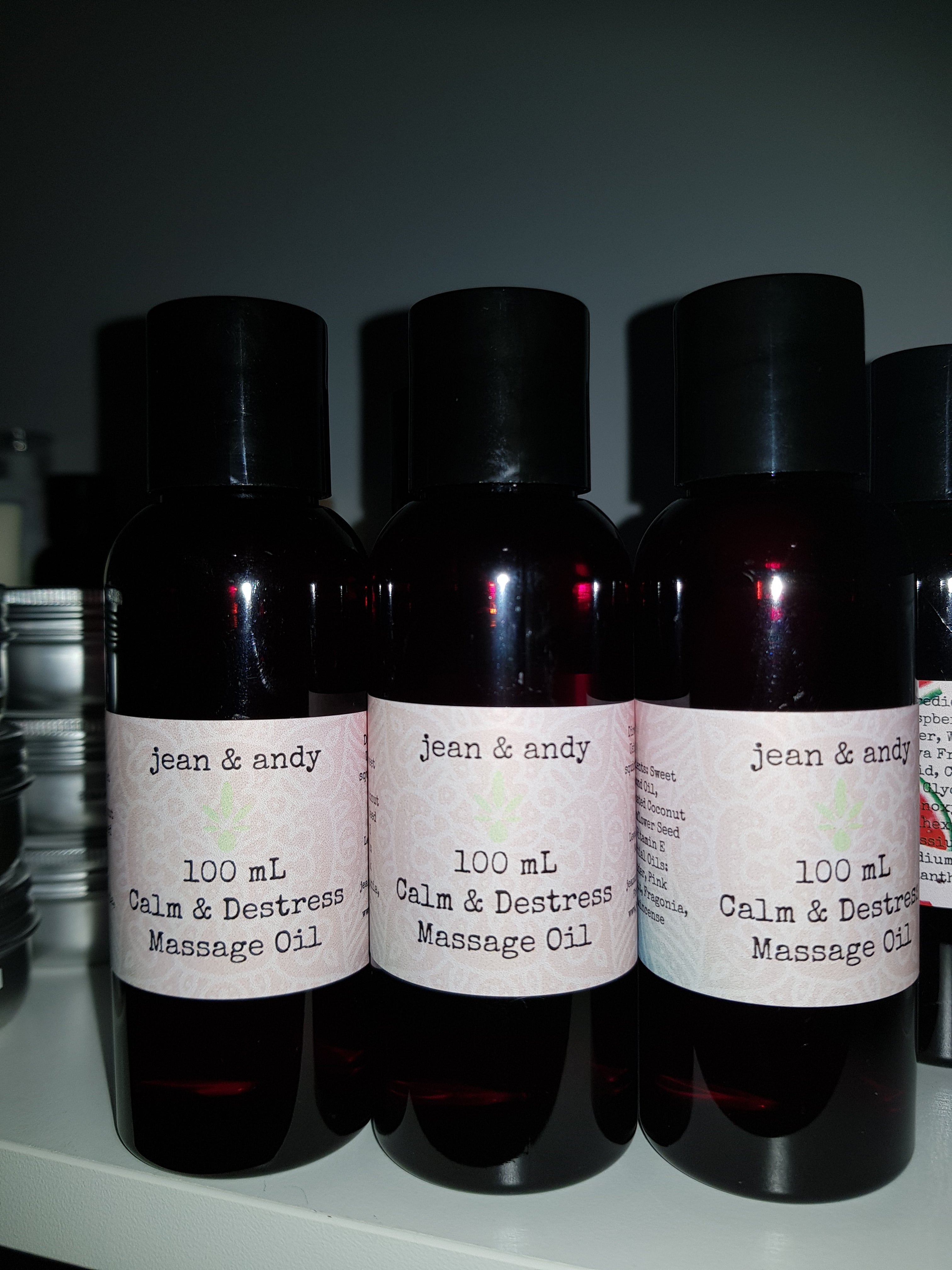 A trio of dark bottles labeled as jean & andy's Calm & Destress Massage Oil, 100 mL each, infused with essential oils for ultimate relaxation, are arranged side by side on a shelf.