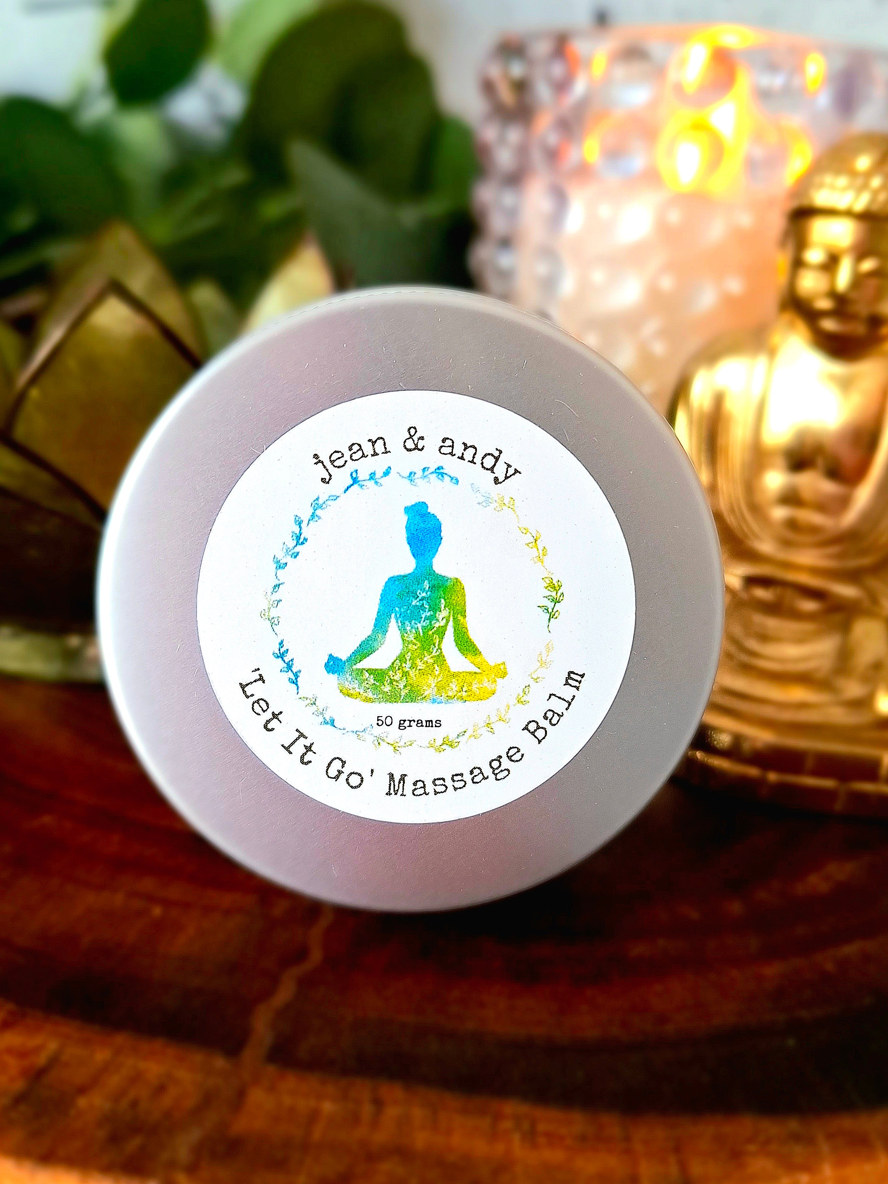 A round tin of Let It Go Massage Balm by jean & andy rests on a wooden surface, surrounded by a glowing candle, a small Buddha statue, and leafy greens. It infuses the space with essential oils, creating a calming experience for the senses.