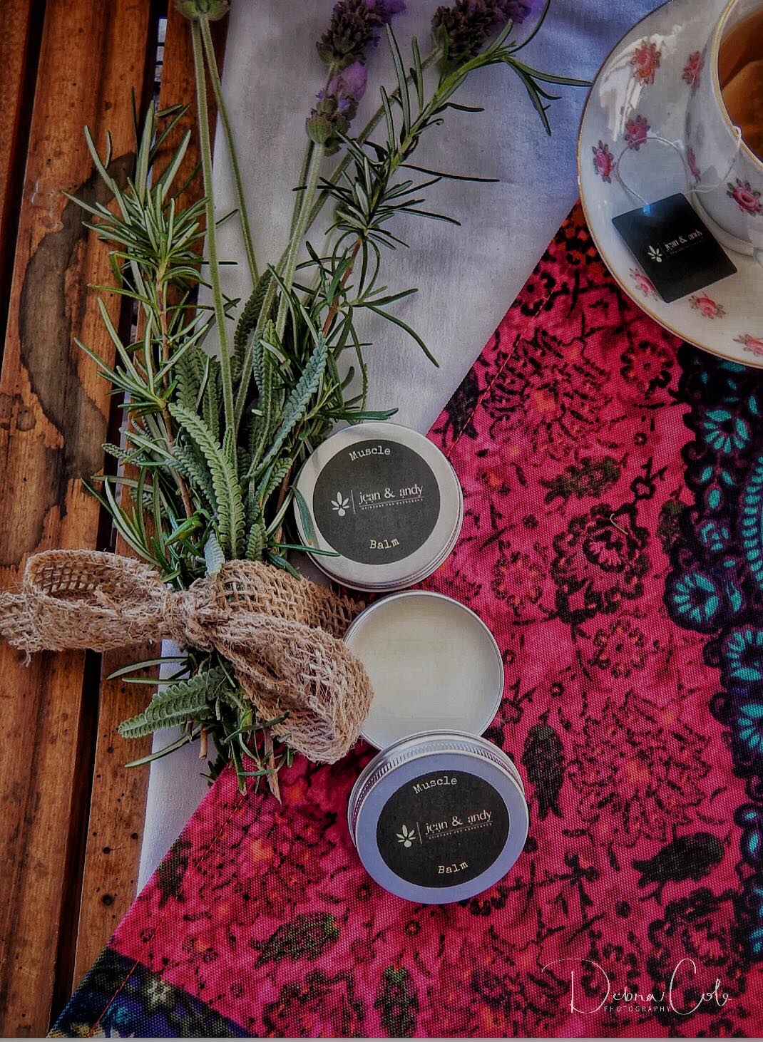 On a wooden table, two tins of jean & andy Muscle Balm sit, one with its lid off, surrounded by sprigs of lavender and greenery tied with burlap. Beneath them lies a vibrant pink patterned fabric, while a teacup holding a teabag rests on the edge. The soothing essential oils in the balm promise relief for sore muscles.