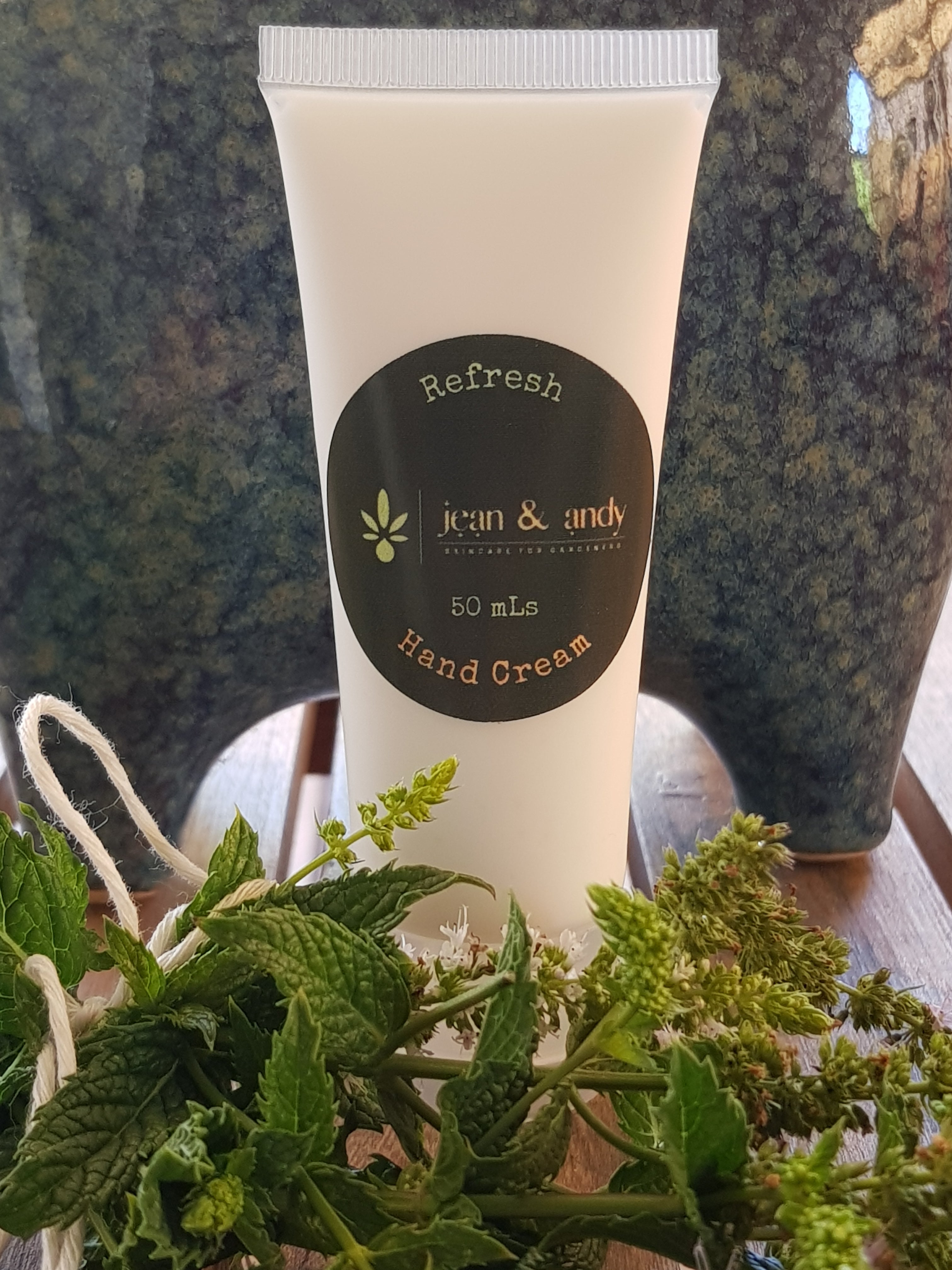A tube of Refresh Hand Cream by jean & andy, infused with invigorating peppermint essential oils, is positioned on a wooden surface. Fresh green herbs are arranged in front of it, with a ceramic pot seen in the background.