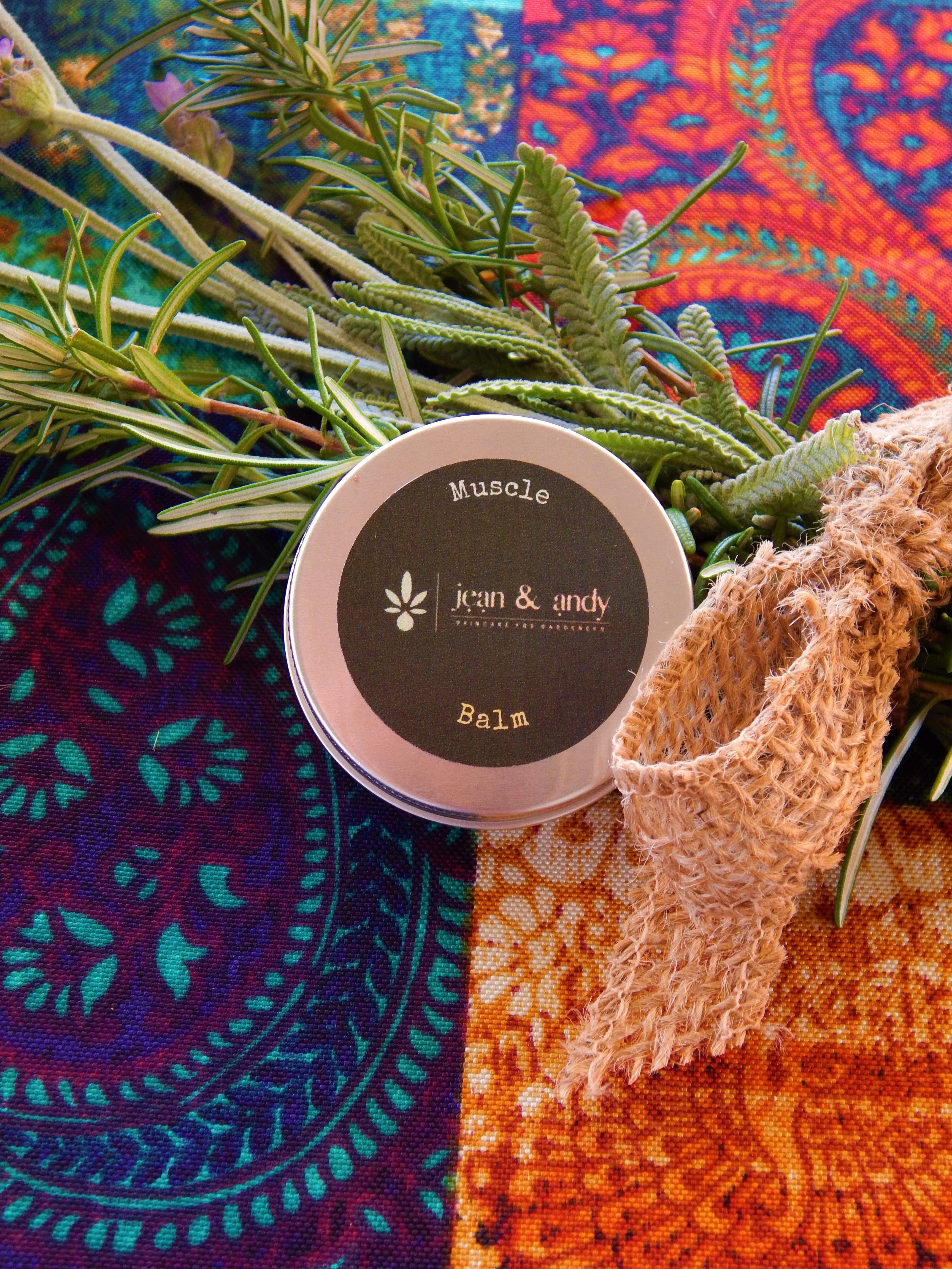 A tin of Muscle Balm from jean & andy sits on a vibrant, patterned fabric. It is enhanced with essential oils for pain relief. Sprigs of rosemary and a burlap ribbon add to the rustic, herbal look.